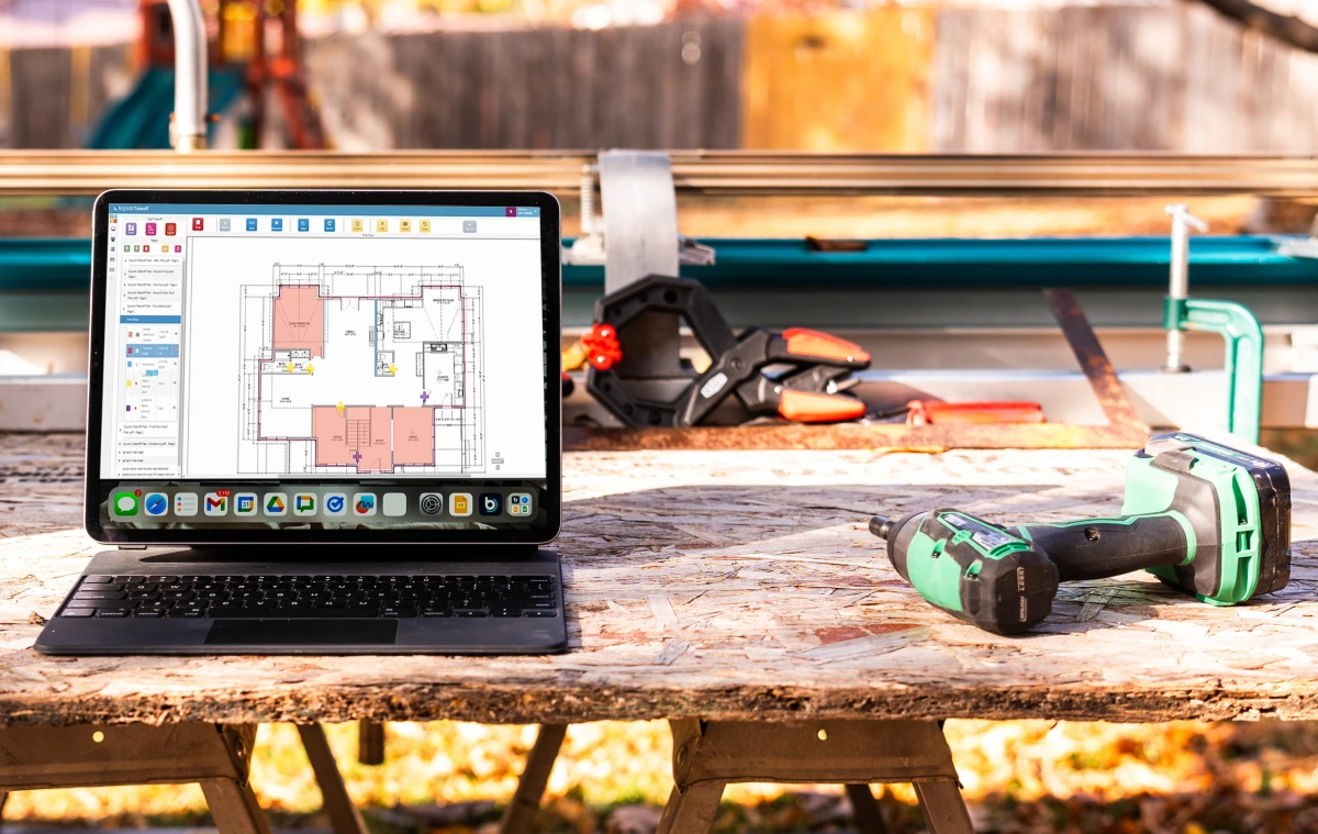 Laptop displaying digital house plans on a construction workbench next to a cordless drill, with building tools and materials in the background.