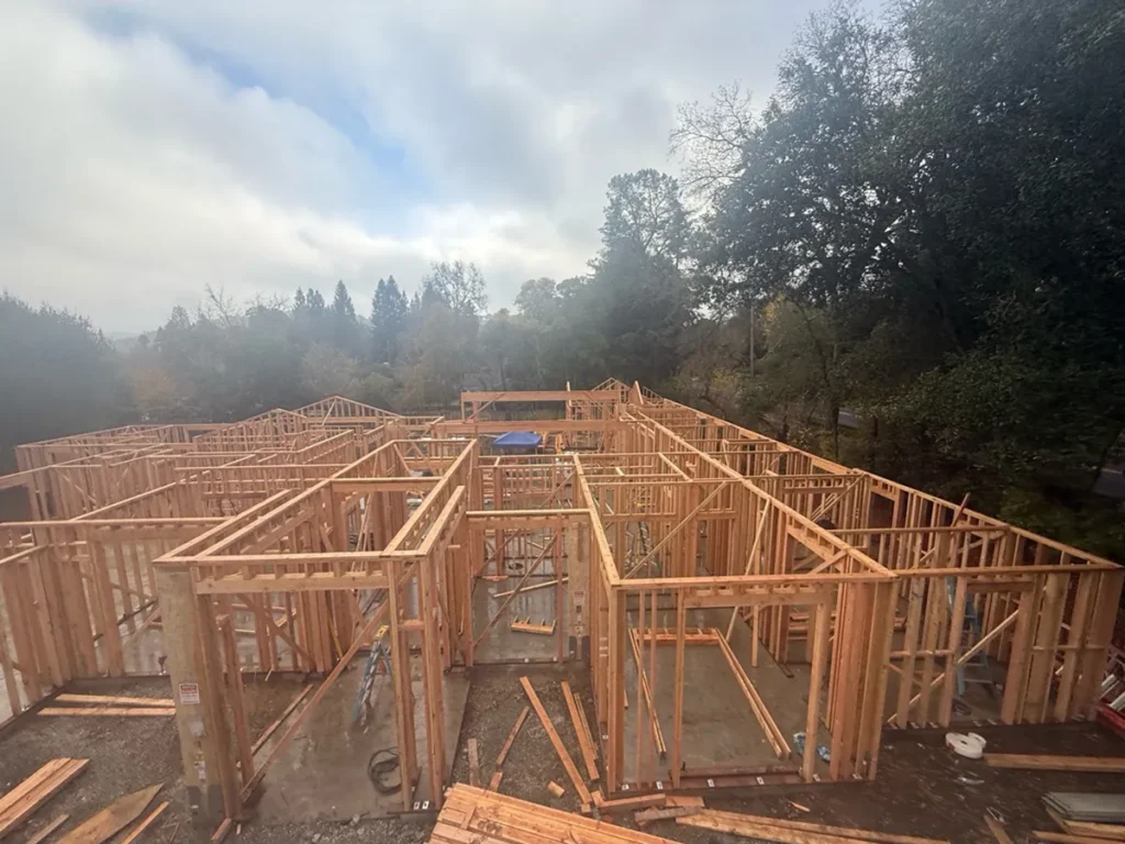Wood framing in progress at a residential construction site, showing wall studs and room layouts before roofing and exterior finishes are installed.